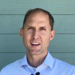 Man in white shirt standing in front of a light blue wooden wall, smiling and looking forward.